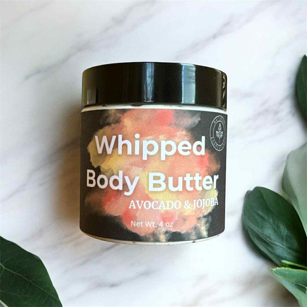 A closed jar of whipped body butter with avocado and jojoba. Set on a piece of marble countertop with leaves framing the photo. 