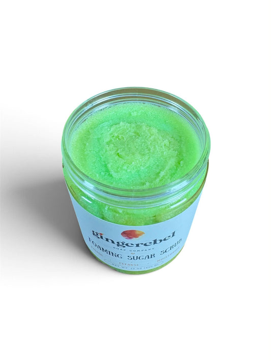 A clear jar of vibrant green flaming sugar scrub with a light blue label, photographed from an overhead angle on a white background.