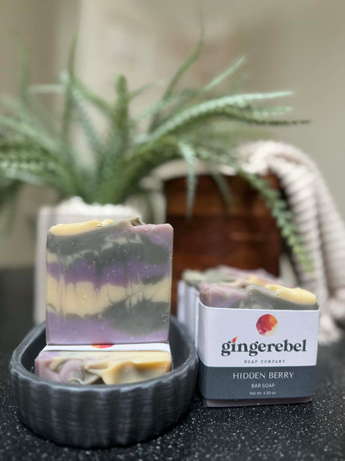 Gingerebel Hidden Berry soap bar with packaging on a dark surface with a plant in the background