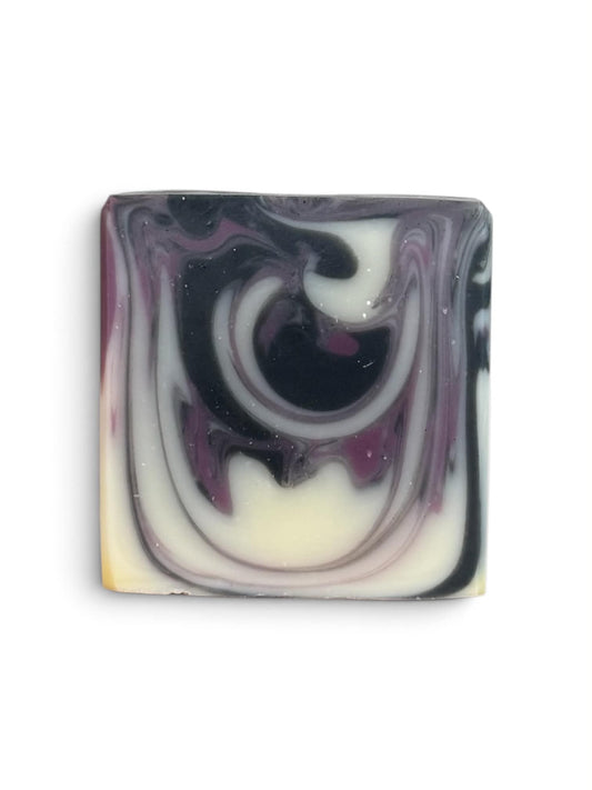 A rectangular bar of handmade soap with a marbled swirl pattern of black, white, and purple, with a small hint of pale yellow at the bottom left corner, set against a plain white background.