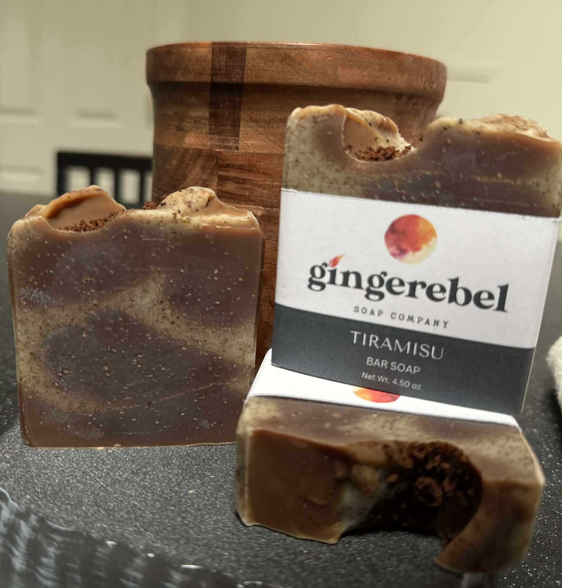 Two bars of Ginger Rebel Tiramisu soap on a dark surface with a wooden container in the background.