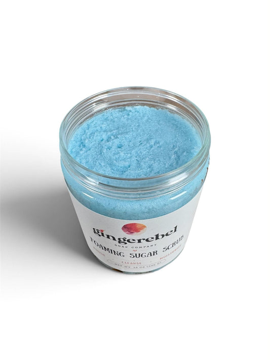 A clear plastic jar filled with light blue foaming sugar scrub, featuring a white label with the brand name 'Gingerebel' and product text, set against a plain white background.