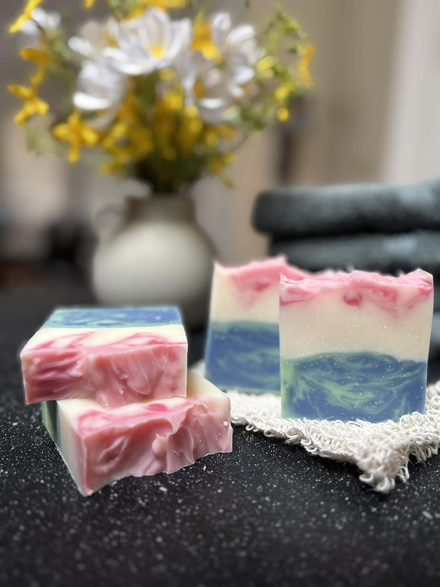 Three bars of soap with a wavy pattern on a dark surface, with a blurred background of flowers and a vase.