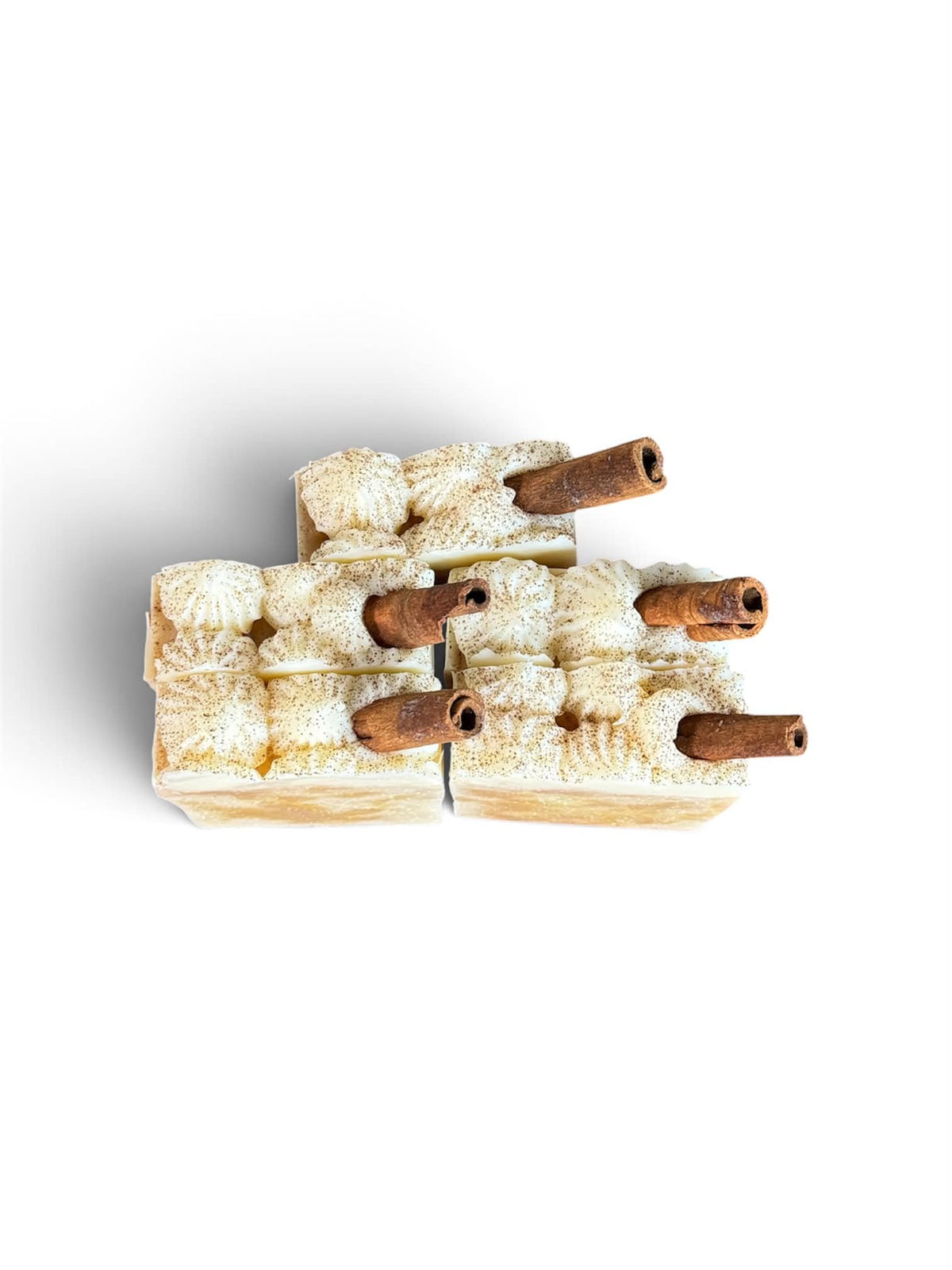 A top-down view of a decorative candle made from layered beige and cream textured wax blocks, each with a small cinnamon stick protruding from the top, set against a plain white background.