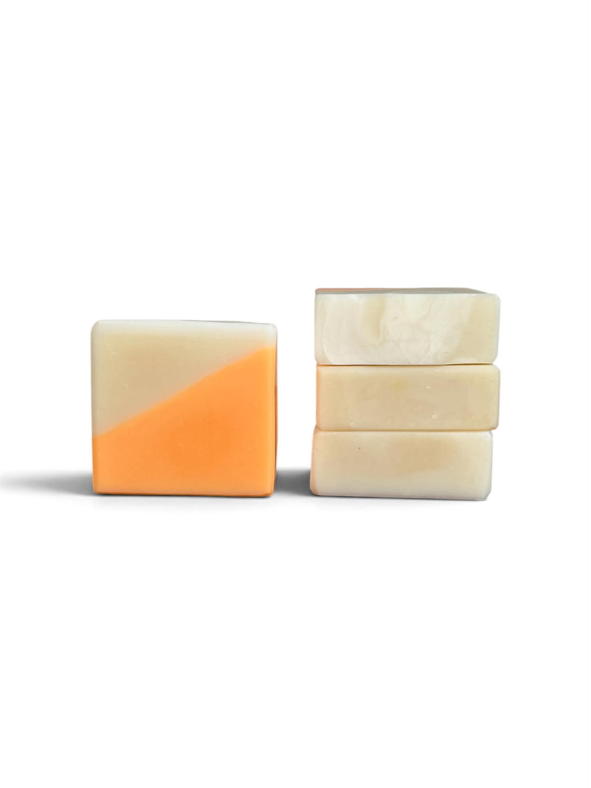 Two rectangular bars of soap on a white background, with one bar showing a cream top half and orange bottom half, and a stack of three cream-colored marbled soap bars.