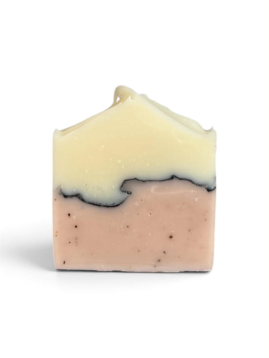 A rectangular bar of handmade soap with a two-tone design, featuring a pale cream-colored top layer and a light pink bottom layer with small dark specks, set against a plain white background.