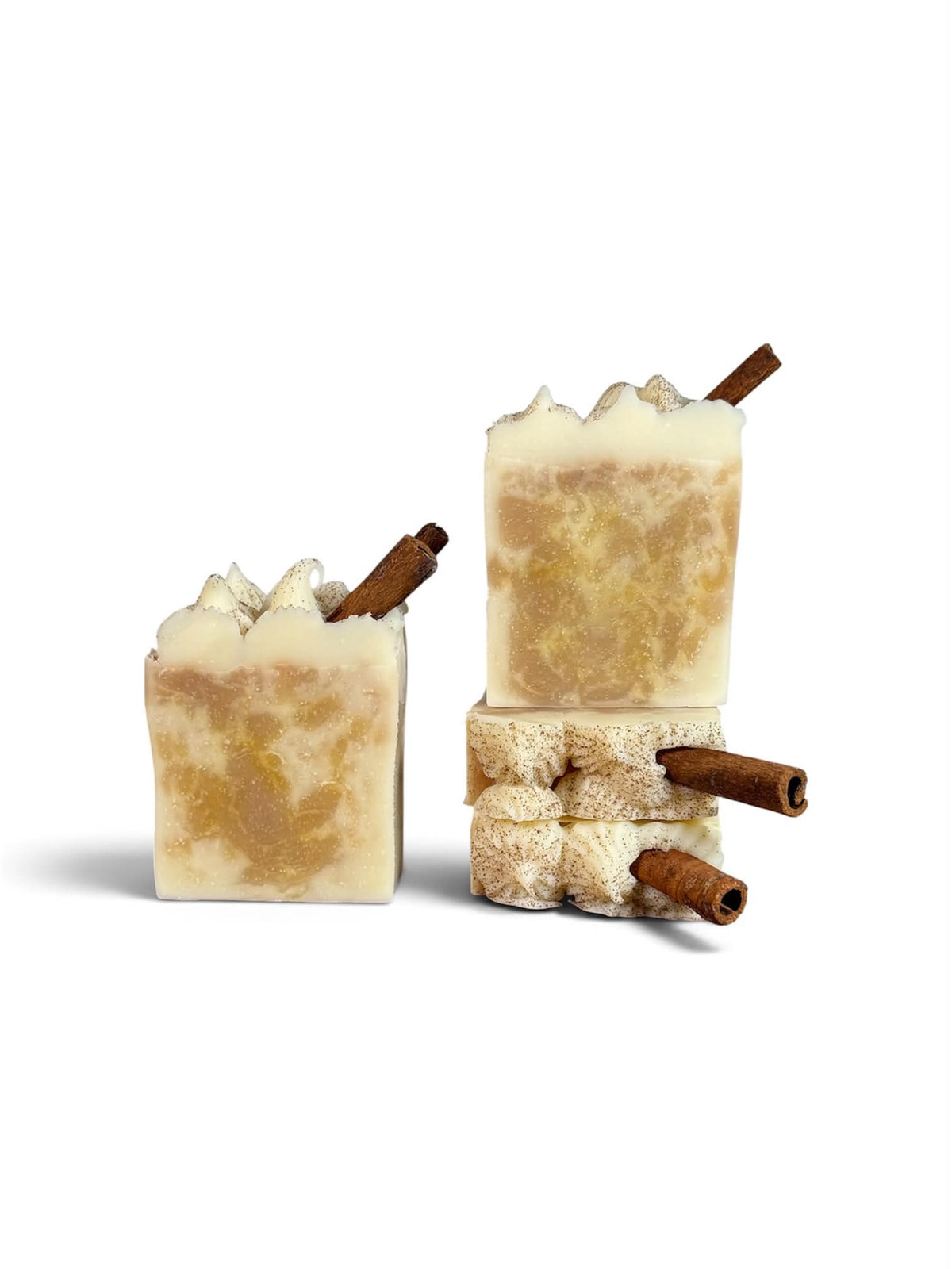 Three handmade rectangular bars of soap with a marbled cream and golden-brown pattern, each topped with whipped cream and decorated with a cinnamon stick, arranged on a white background.