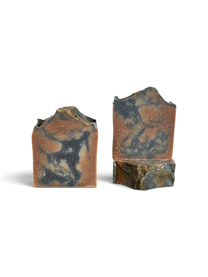Two rustic, hand-cut soap bars with a marbled pattern of terracotta orange, black, and brown, displayed on a clean white background.
