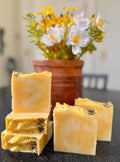 Yellow soap bars with specks on a dark surface, with a pot of flowers in the background.