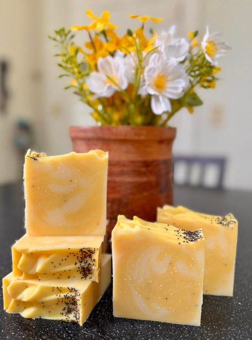 Yellow soap bars with specks on a dark surface, with a pot of flowers in the background.