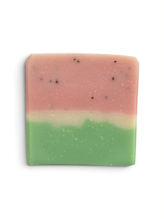 A rectangular bar of handmade soap with a two-tone design, featuring a pink top half and a green bottom half, separated by a thin pale cream band. Small black specks are visible throughout the soap, and it is photographed on a clean white background.